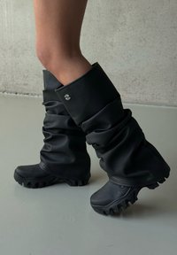 Jambes portant des bottes noires surdimensionnées et décontractées avec des semelles épaisses et robustes, debout sur un sol gris lisse contre un mur texturé.