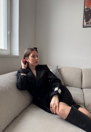 Blazer negro con acentos blancos, sentado en un sofá gris claro. Largo corto, tejido texturizado, combinado con botas altas negras.