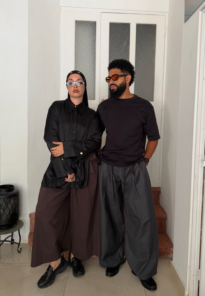 Deux personnes portant des vêtements amples et sombres ainsi que des lunettes de soleil se tiennent à l'intérieur près d'une porte blanche avec des panneaux de verre dépoli.