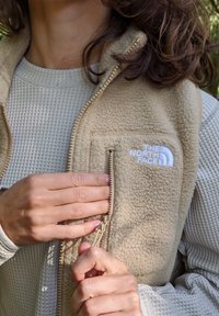 Beige Fleece-Weste mit einer zipfbaren Brusttasche. Das North Face-Logo ist in weiß gestickt. Darunter befindet sich ein strukturiertes, hellgraues Oberteil.