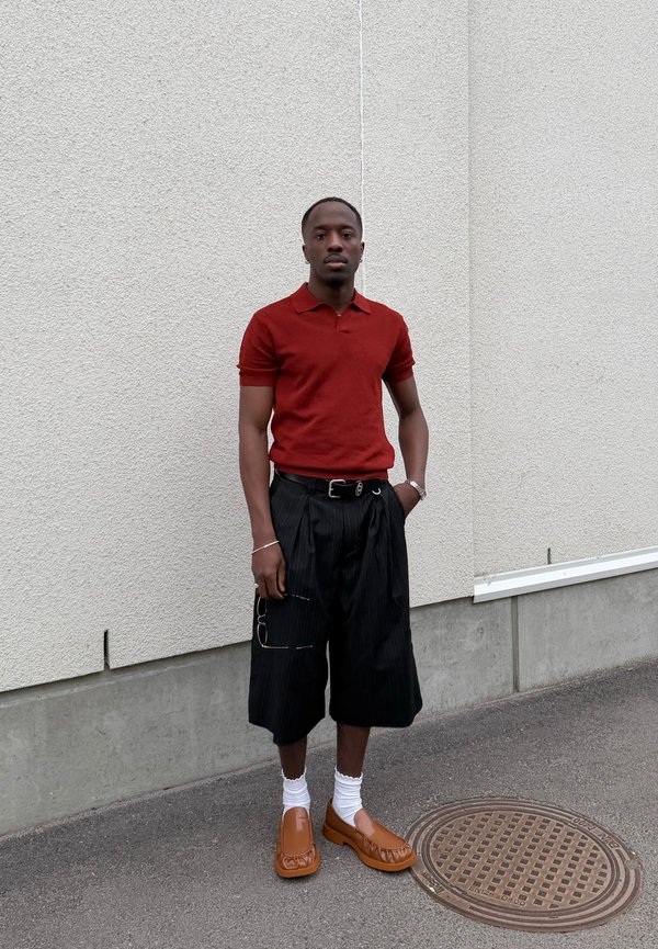 Bărbat purtând un tricou polo roșu, pantaloni scurți negri largi, șosete albe și mocasini maro, stă lângă un perete alb texturat pe o paviment gri.
