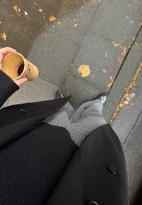 Cappotto nero, maglione a coste nero, pantaloni grigi a righe, scarpe nere lucide; tiene in mano una tazza con una bevanda marrone su un marciapiede bagnato.