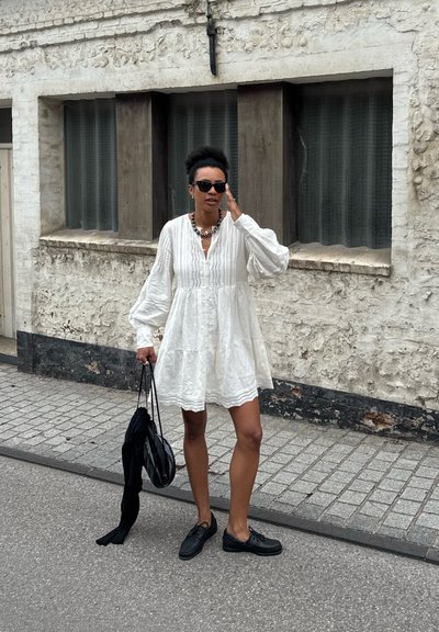Un vestido de lino blanco con mangas abullonadas y un diseño de capas. Zapatos negros y un bolso negro, con gafas de sol y un collar llamativo como acentos.