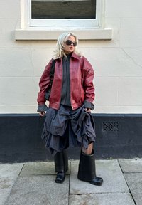 Crvena kožna jakna, sivi pleteni džemper, siva naborana suknja, crne mrežaste čarape i velike crne čizme. Stoji naslonjena na svijetli zid.