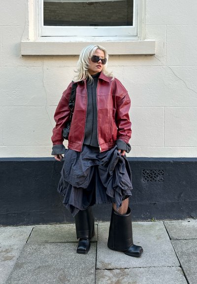 Chaqueta de cuero roja, suéter de punto gris, falda gris con volantes, medias de red negras y botas negras de gran tamaño. De pie contra una pared clara.
