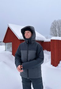 Gevoerde jas in grijs en zwart, met een hoge kraag en capuchon. Gladde textuur, rits aan de voorkant en zijzakken. Sneeuwachtige achtergrond.