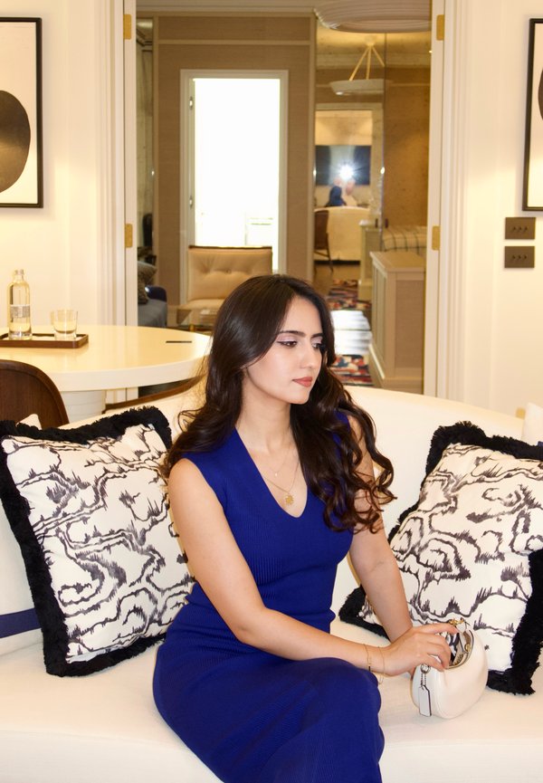 Woman in a blue dress sitting on a white couch with patterned black and white pillows, holding a small white handbag inside a modern room.