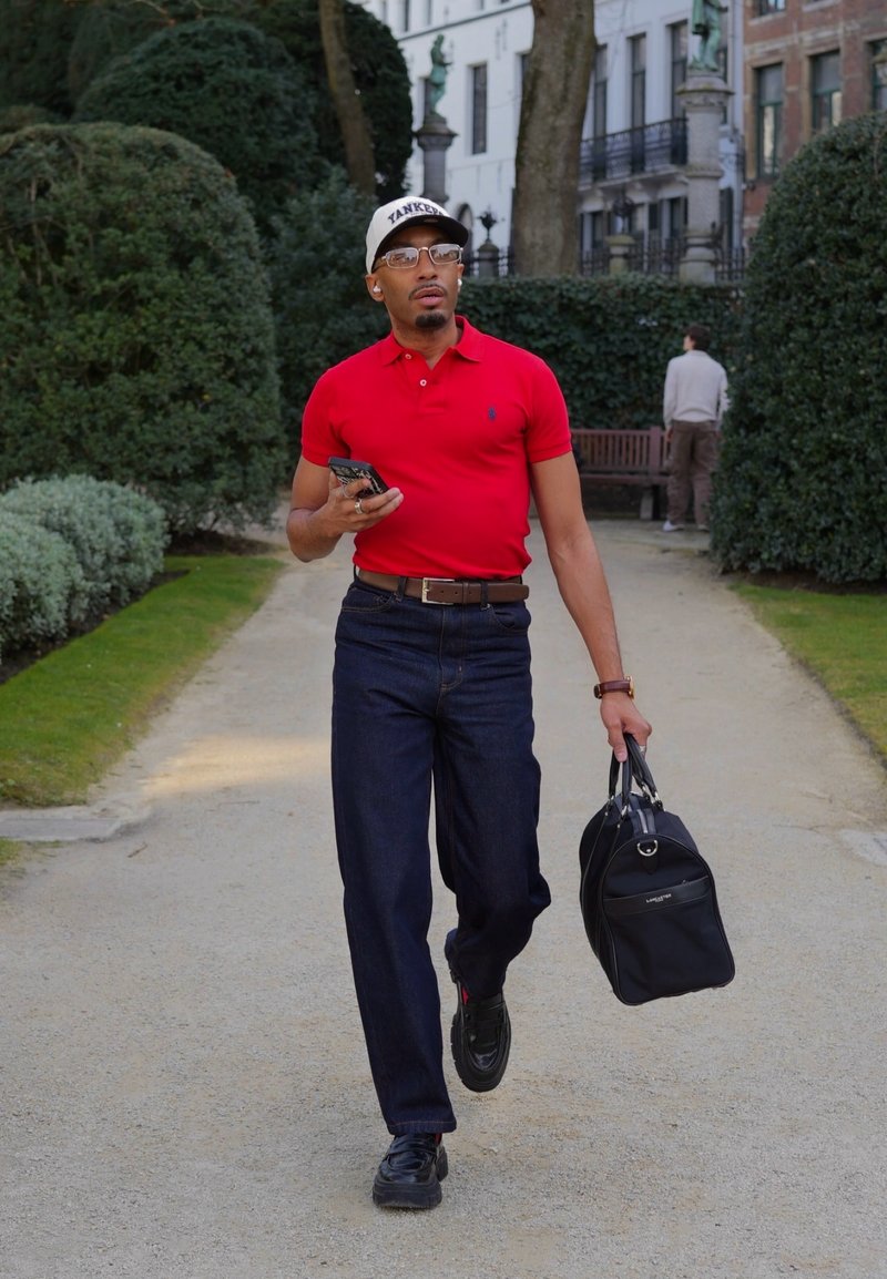 Homme en polo rouge, jean foncé et casquette marchant sur un chemin de parc, tenant un téléphone et un sac noir, entouré de verdure et de bâtiments.