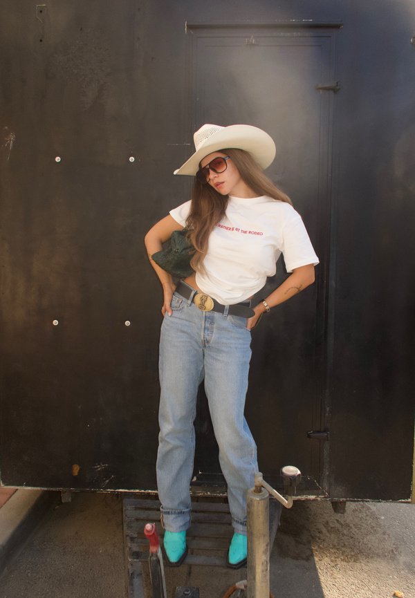 Noor naine, kes kannab valget cowboy-mütsi, päikeseprille, valget t-särki, siniseid teksapükse ja türkiissiniseid saapaid, seisab musta seina ees.