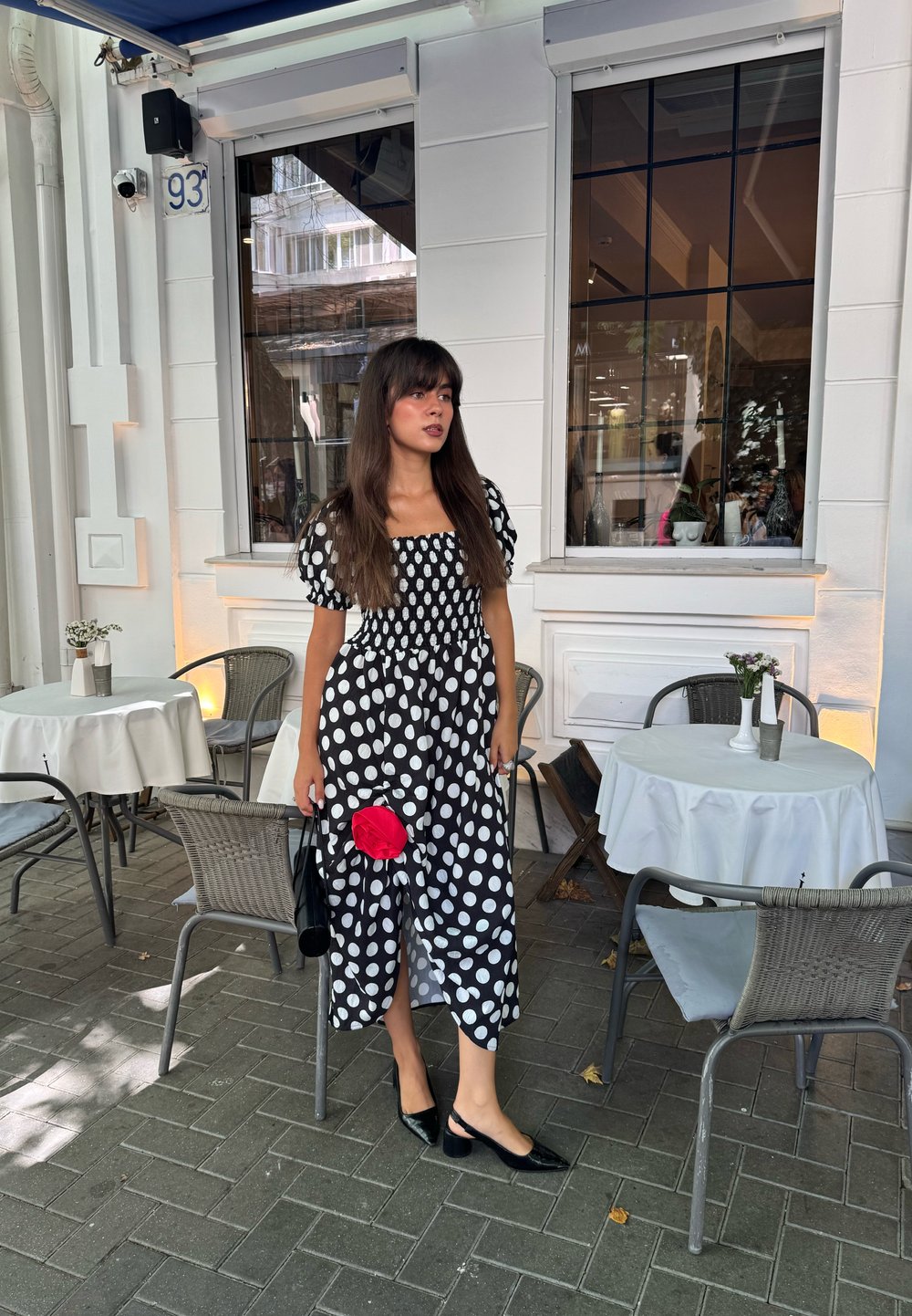 Donna in un vestito a pois bianco e nero tiene in mano un fiore rosso e si trova accanto ai tavoli di un caffè all'aperto con tovaglie bianche.