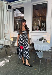 Donna in un vestito a pois bianco e nero tiene in mano un fiore rosso e si trova accanto ai tavoli di un caffè all'aperto con tovaglie bianche.