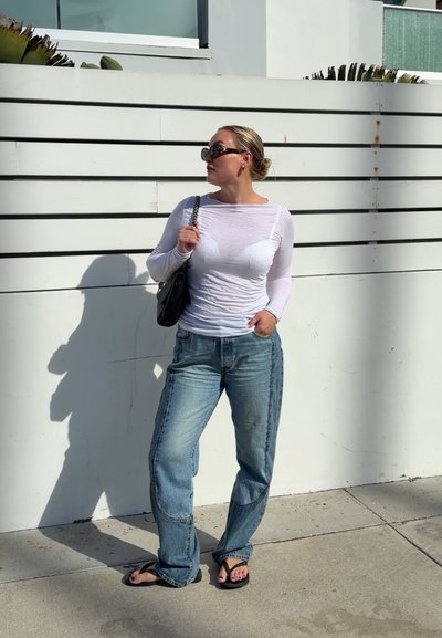 Mujer con gafas de sol, camiseta blanca de manga larga, vaqueros azules y chanclas, sosteniendo un bolso negro de hombro, de pie junto a una valla blanca.