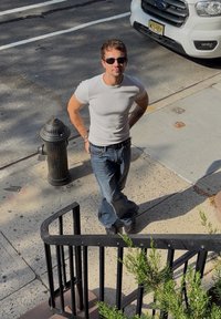 Tricou alb, blugi cu o croială lejeră, ochelari de soare negri, stând lângă o hidrantă de pompieri neagră și o balustradă curbată neagră.