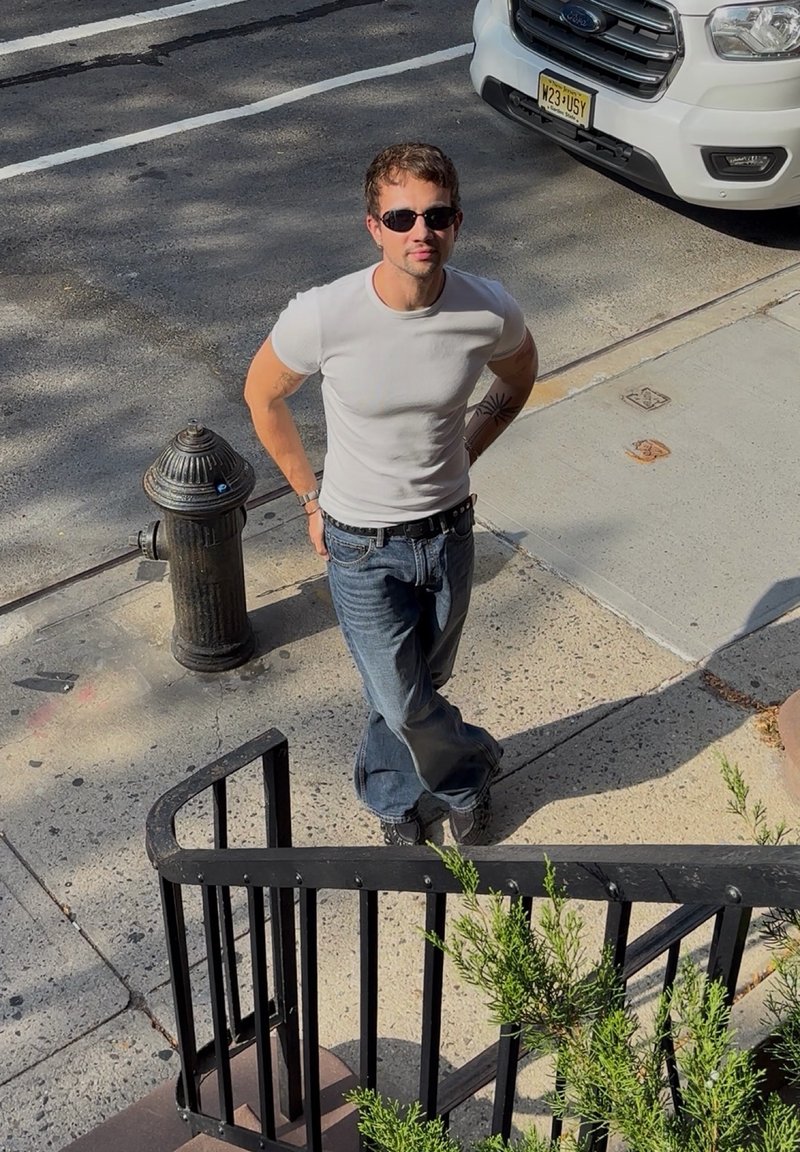 T-shirt bianco, jeans blu con una vestibilità comoda, occhiali da sole neri, in piedi vicino a un idrante nero e a una ringhiera curva nera.
