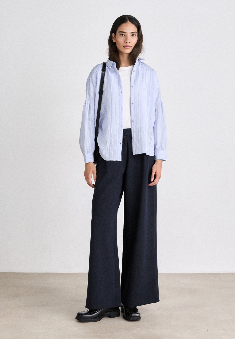 Svetlomodrá pruhovaná nadmerná košeľa, biela tričko pod ňou, skombinovaná s vysokými nohavicami s širokými nohavicami v námorníckej modrej farbe a čiernymi platformovými topánkami.