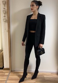 Femme aux cheveux en chignon portant un blazer noir, un pantalon ajusté, des bottes et un crop top, tenant un petit sac à main noir, debout à l'intérieur sur un sol en bois.