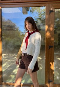 Gebreide crèmekleurige trui met een losse pasvorm en een rode sjaal, gecombineerd met donkerbruine suède shorts. Op de achtergrond een houten deur en natuurlijk licht.