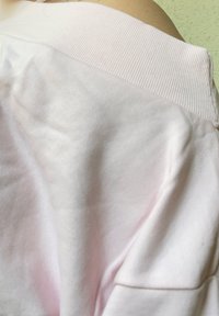 Tecido de algodão rosa claro, suave, com uma gola canelada. Textura lisa, com costuras visíveis e rugas subtis. Corte casual e relaxado.