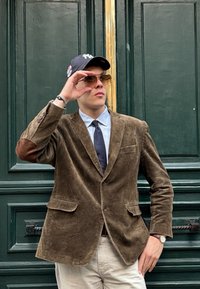 Blazer in corduroy marrone con toppe sui gomiti, camicia button-up azzurra chiara, cravatta scura, occhiali da sole e cappello nero di fronte a una porta verde.