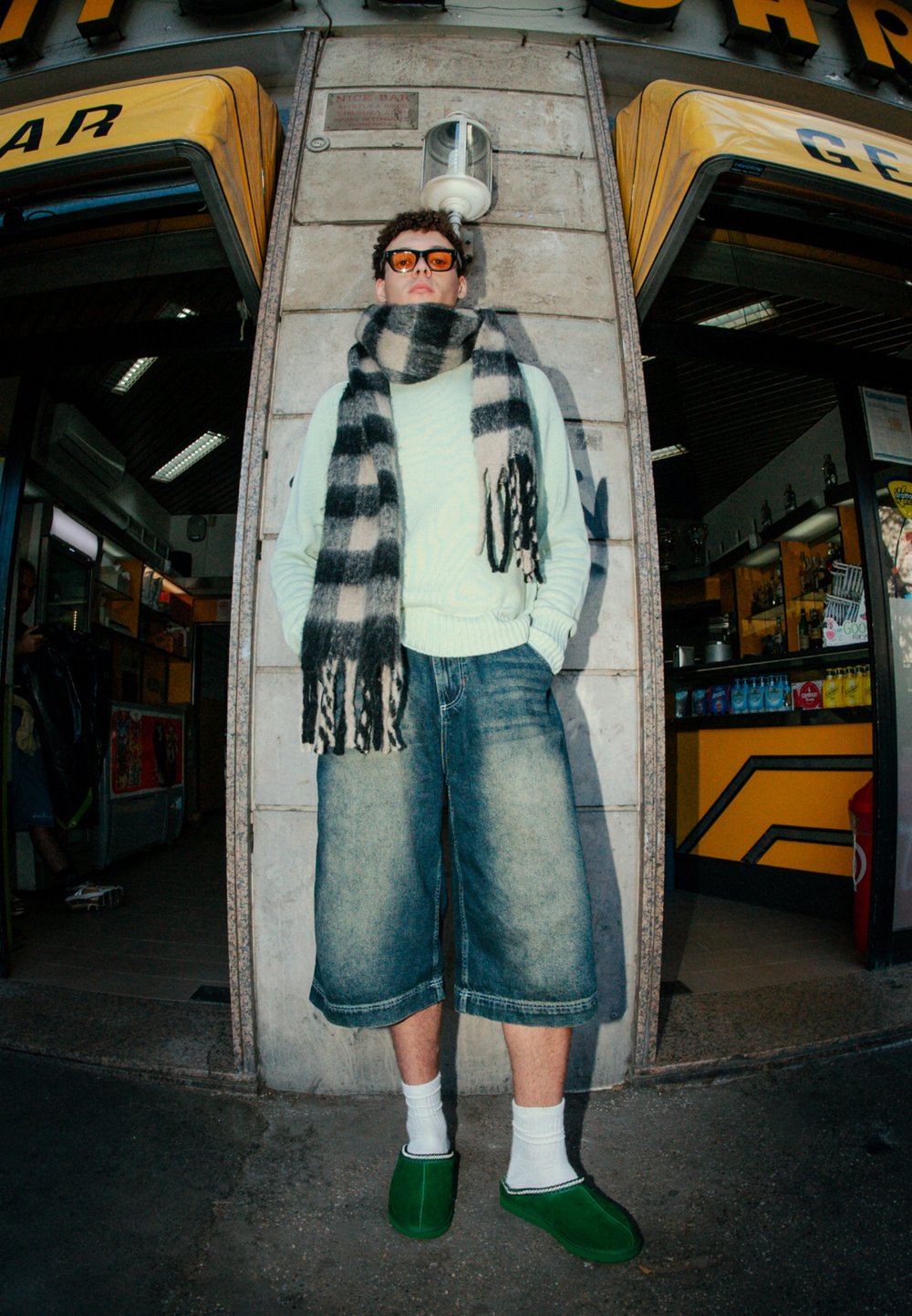 Light green jumper, black and white striped scarf, oversized denim shorts, white socks, and green footwear in an urban setting.