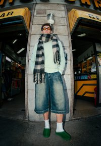 Light green jumper, black and white striped scarf, oversized denim shorts, white socks, and green footwear in an urban setting.