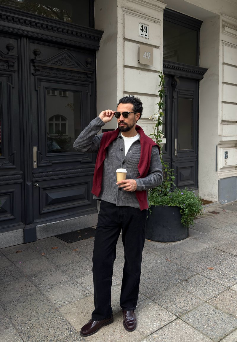 Hombre con gafas de sol, cárdigan gris y suéter rojo sostiene una taza de café frente a un edificio con puertas negras y el número 49.