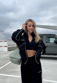 Sort lynlåstrøje med hvide detaljer og cropped top, kombineret med sorte bukser. Grøn skuldertaske og gråt køretøj i baggrunden.