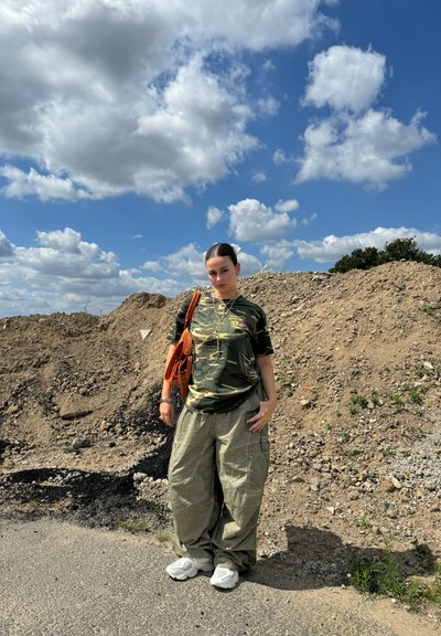 Camiseta de camuflaje, pantalones cargo holgados oliva, zapatillas blancas y una bolsa naranja. Fondo texturizado de tierra y cielo azul con nubes.