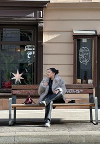 Model, kas sēž uz koka sola, ģērbusies pelēkā mākslīgā ādas mētelī un melnās ādas biksēs, blakus viņai burgundijas krāsas rokas soma. Veikala logā izvietoti augi.