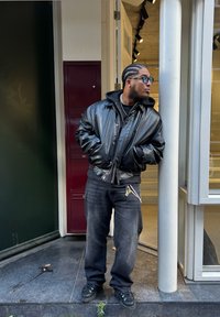 Must musta bomber-jakk musta nahast, millel on karvaga vooderdatud krae, koos laste mustade teksade ja mustade tossudega. Seisab hoone sissepääsu lähedal.
