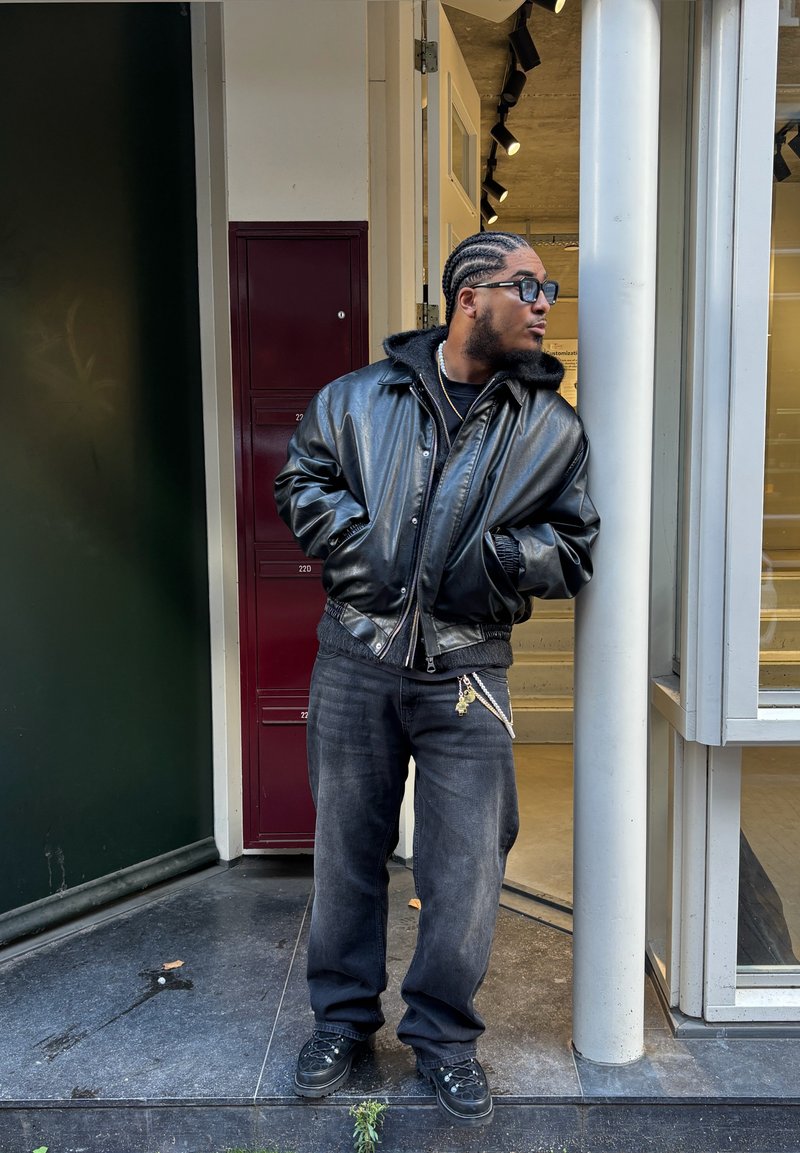 Chaqueta bomber de cuero negra con cuello forrado de piel, combinada con vaqueros negros holgados y zapatillas negras. De pie cerca de la entrada de un edificio.