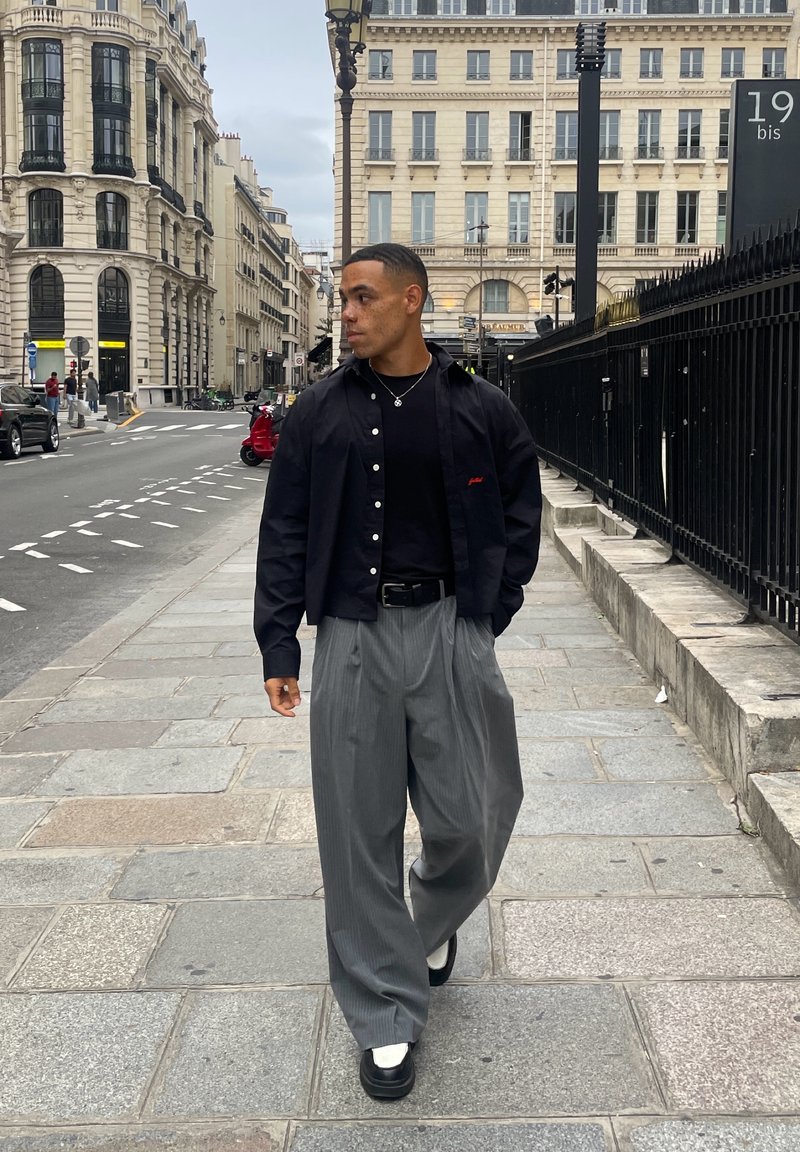 Black button-up shirt over a black t-shirt, gray pleated trousers, and black shoes with white soles. Urban street setting with buildings.
