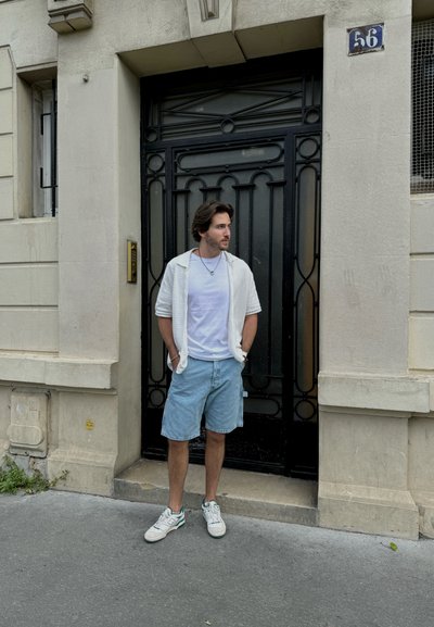 Una persona lleva un suéter de punto blanco sobre una camiseta blanca y pantalones cortos de denim, de pie frente a una puerta decorativa negra con forja.