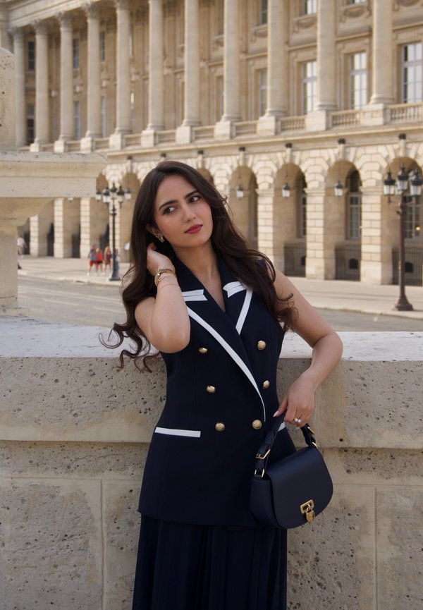 Mujer con cabello largo y oscuro, vestida con un vestido abrigo sin mangas en blanco y negro, sosteniendo un bolso negro, está junto a un muro de piedra con un edificio histórico de fondo.