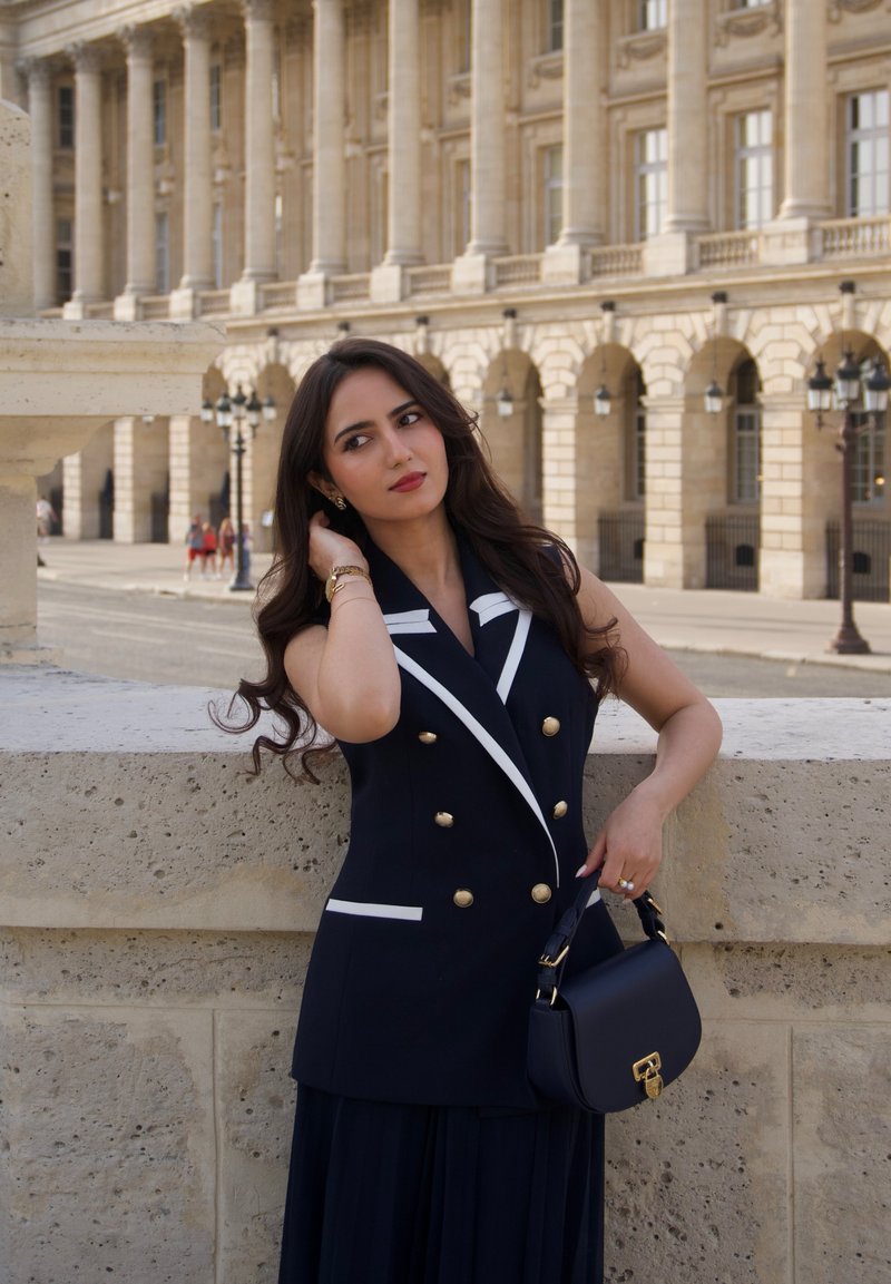 Woman with long dark hair in black and white sleeveless coat dress, holding black handbag, stands by stone wall with historic building in background.