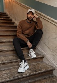 Hombre sentado en las escaleras, vistiendo un suéter marrón y pantalones negros, con zapatillas blancas que tienen detalles en gris y cordones texturizados.