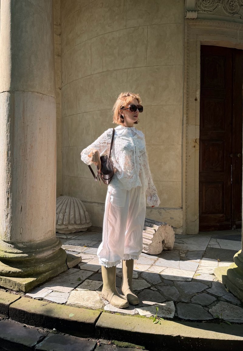 Femme portant un chemisier en dentelle blanche, un pantalon court blanc, des bottes beiges, des lunettes de soleil, et tenant un sac marron, debout près de colonnes et d'un mur en pierre anciens.