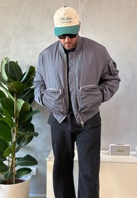 Giacca bomber grigia con trama trapuntata, dotata di due tasche frontali con zip e una tasca laterale, abbinata a pantaloni neri e un berretto.