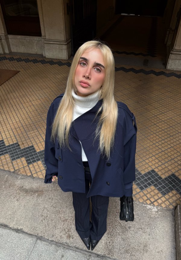 Donna bionda con cappotto blu navy e dolcevita bianco in piedi su un pavimento piastrellato, che tiene una borsa nera e guarda verso l'alto verso la fotocamera.