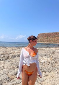 Bikini s oranžovými a žltými vzormi, doplnený voľnou bielou košou. Prostredie obsahuje skalnatý terén pri mori a modrú oblohu.