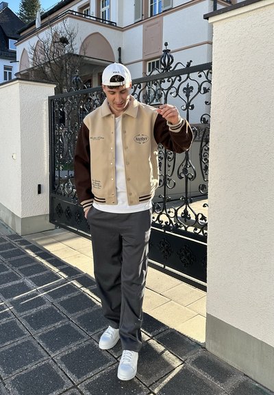 Chaqueta varsity beige y marrón con detalles bordados, camiseta blanca, pantalones grises y zapatillas blancas, de pie sobre un camino pavimentado.