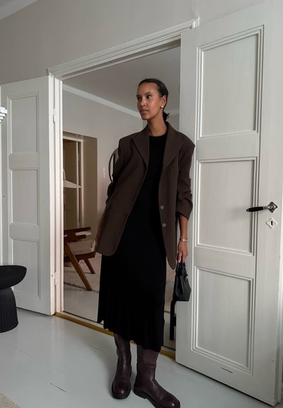 Blazer marrón de gran tamaño sobre un vestido negro de canalé, combinado con botas burdeos altas hasta la rodilla. Sostenido un pequeño bolso negro, de pie en un umbral.
