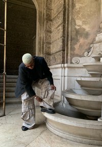 Una figura regola dei pantaloni cargo larghi e chiari, dotati di molte tasche, abbinati a un blazer scuro e scarpe nere, vicino a una fontana in marmo.