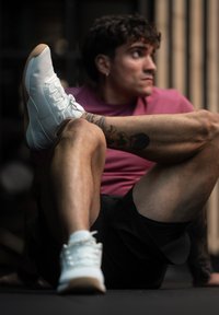 Bílé sportovní boty s texturovaným síťovaným svrškem a lehkou gumovou podrážkou, kombinované s černými šortkami a růžovým tričkem, zdůrazněné viditelnými tetováními.