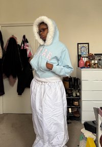 Άτομο που φοράει ανοιχτό μπλε μπουφάν με κουκούλα και γούνα στις άκρες και γυαλιά, στέκεται μέσα σε κτίριο κοντά σε πόρτα και έπιπλο με παπούτσια και διακοσμητικά.