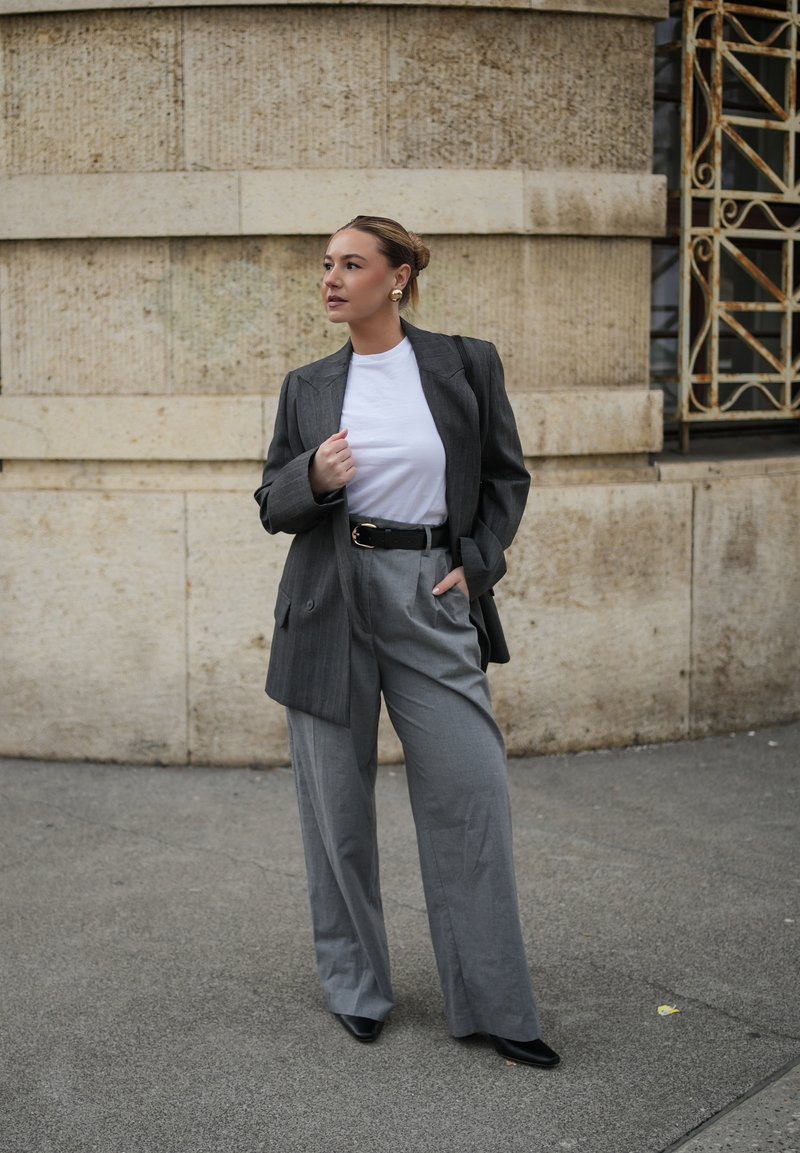 Γυναίκα με γκρι σακάκι, λευκό πουκάμισο, φαρδιά γκρι παντελόνια και μαύρα παπούτσια στέκεται δίπλα σε πέτρινο τοίχο, κοιτάζοντας προς τα πλάγια.