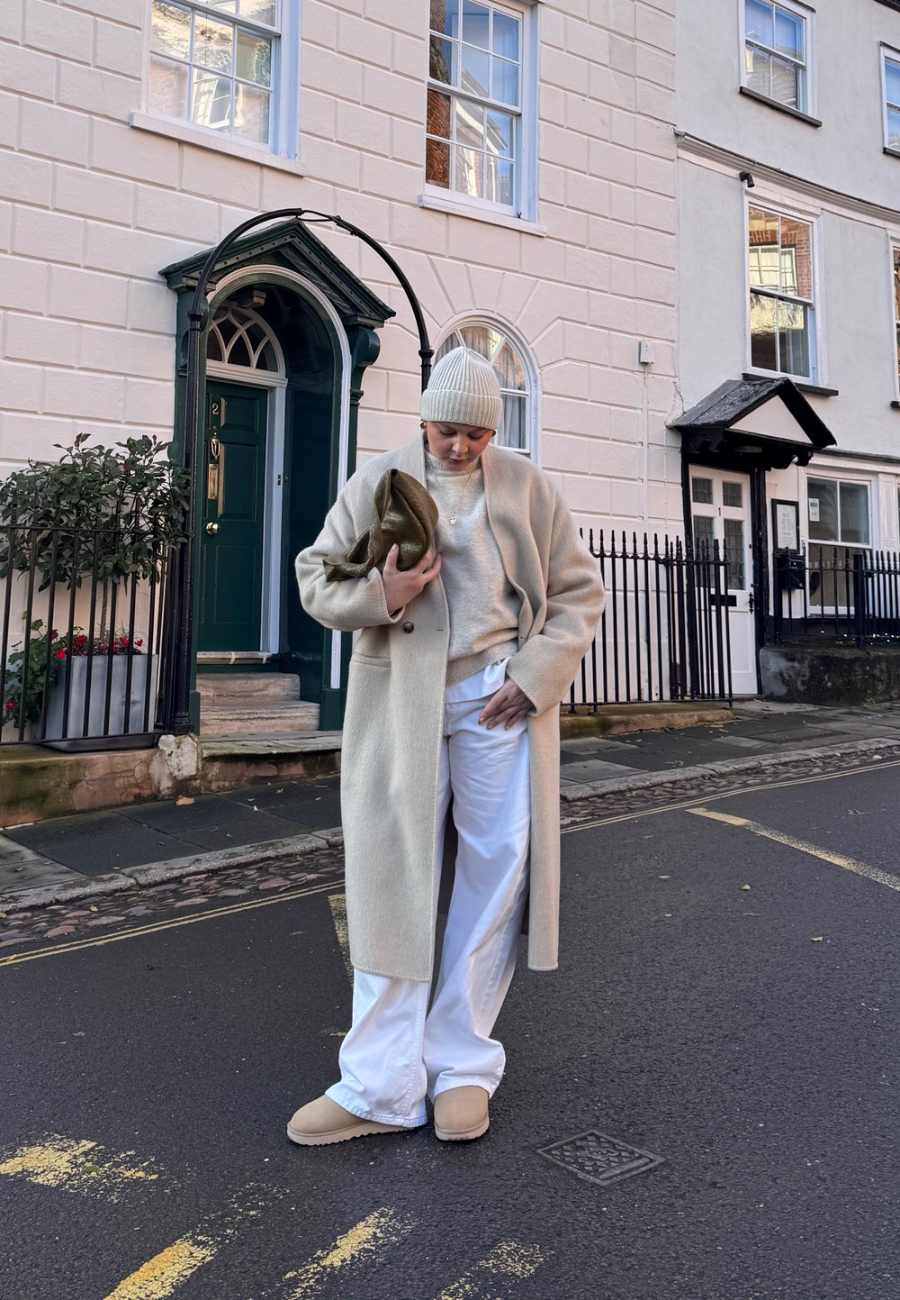 Femme en manteau beige, pantalon blanc et bonnet en tricot se tenant dans la rue avec un sac vert olive, devant un bâtiment couleur crème et une porte verte.