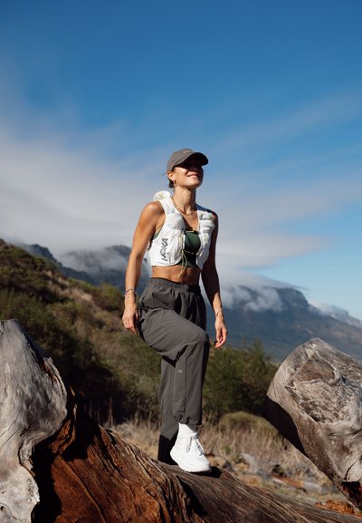 Mujer haciendo senderismo al aire libre sobre un gran tronco, con gorra, chaleco blanco, pantalones oscuros y zapatillas blancas, con montañas y cielo azul al fondo.