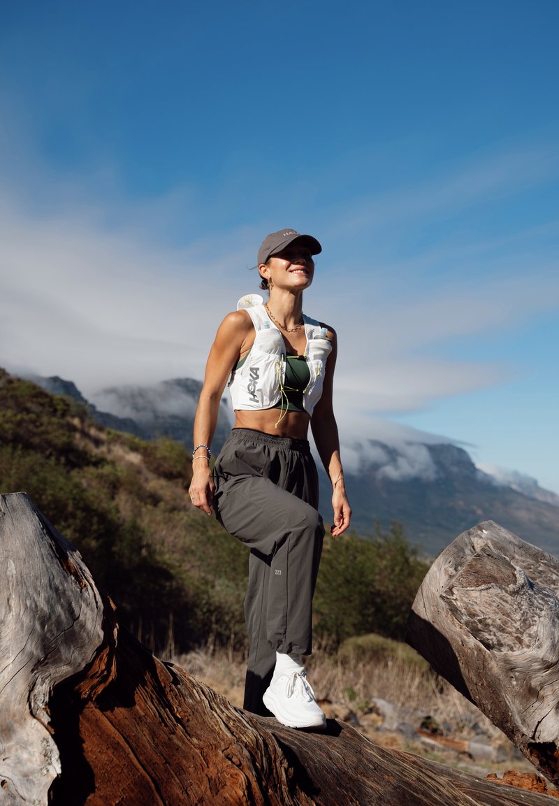 Femme en randonnée à l’extérieur sur une grosse bûche, portant une casquette, un gilet blanc, un pantalon sombre et des baskets blanches, avec des montagnes et un ciel bleu en arrière-plan.