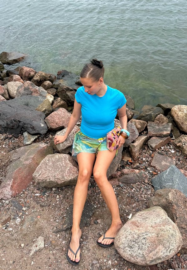 Žena v modrej košeli a šortkách sediaca na skalách pri vode, drží farebnú kabelku, má obuté žabky a vlasy zviazané v drdole.